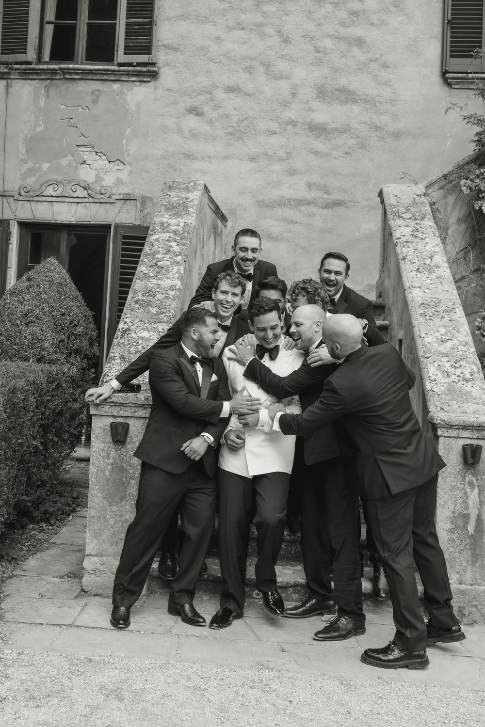 Groom surrounded by groomsmen in black tuxedos, laughing and celebrating on stone steps of a historic venue.