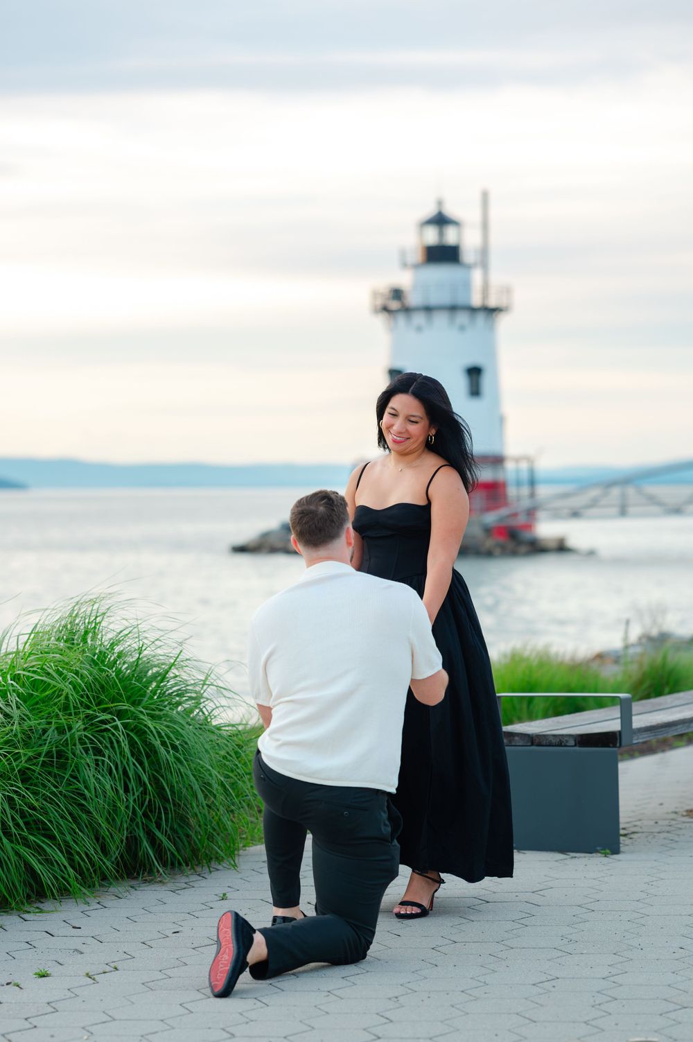 Man proposing on one knee to woman in black dress near waterfront lighthouse.