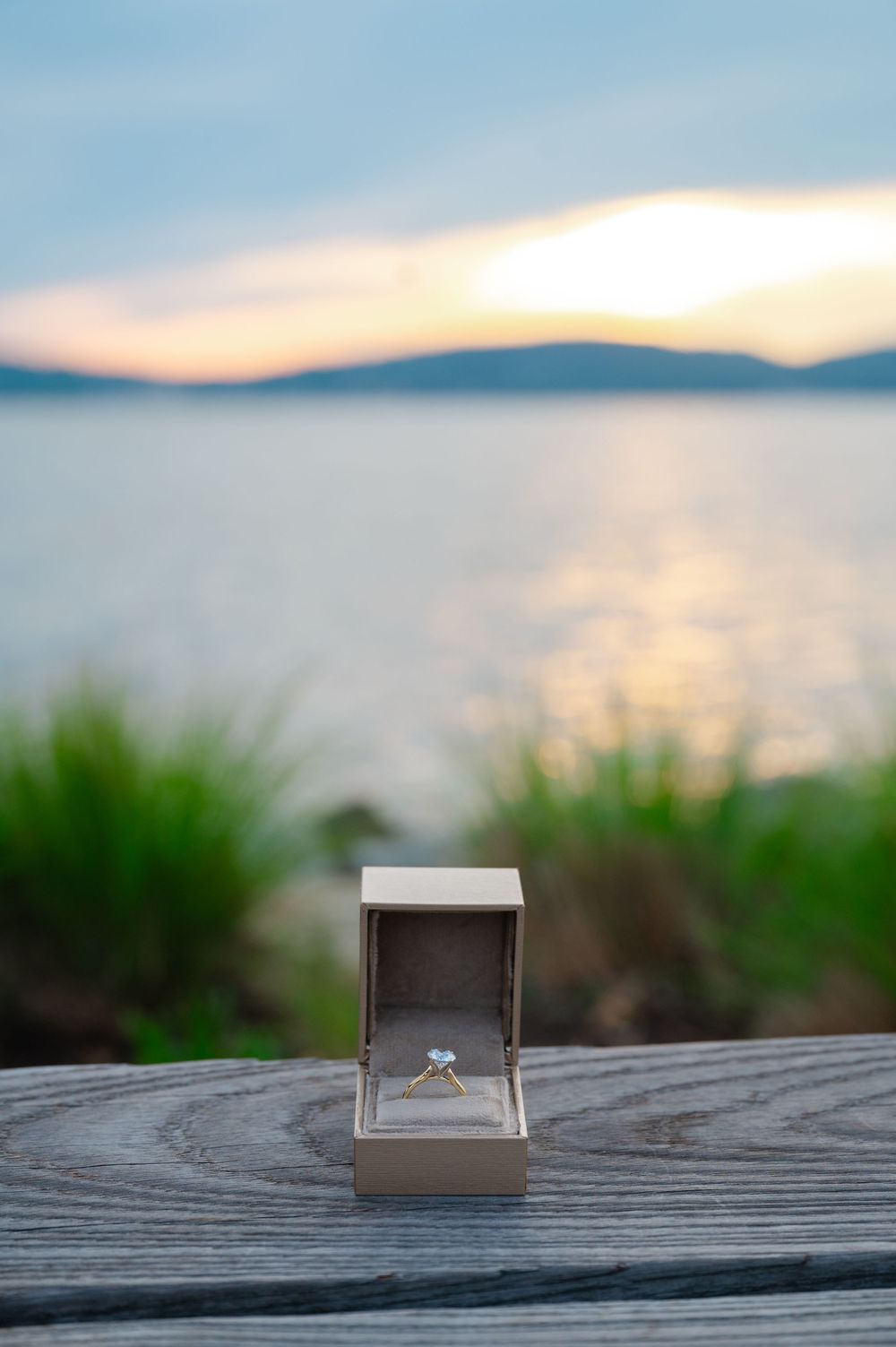 Diamond engagement ring in open box on wooden surface with sunset lake view in background.