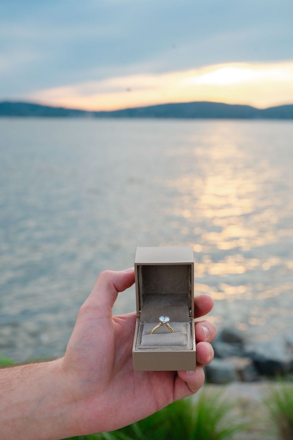 Hand holding open ring box with diamond engagement ring at sunset over calm water with hills in background.