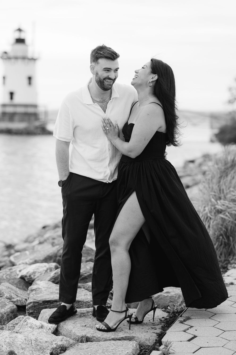 Black and white photo of laughing couple near lighthouse on rocky waterfront, woman in black dress.