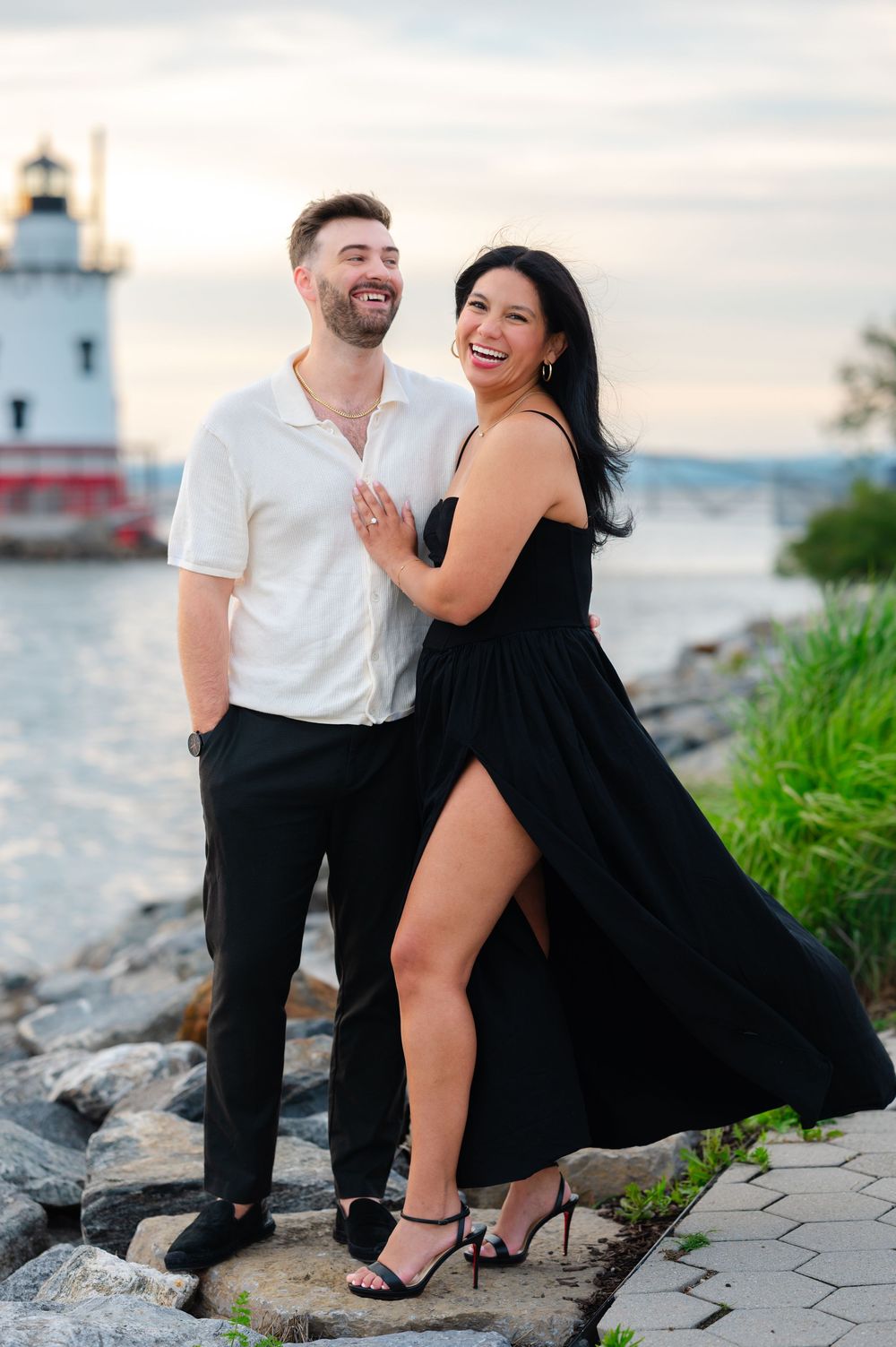Laughing couple posing on rocky shore near lighthouse at sunset, woman in black dress, man in white shirt.