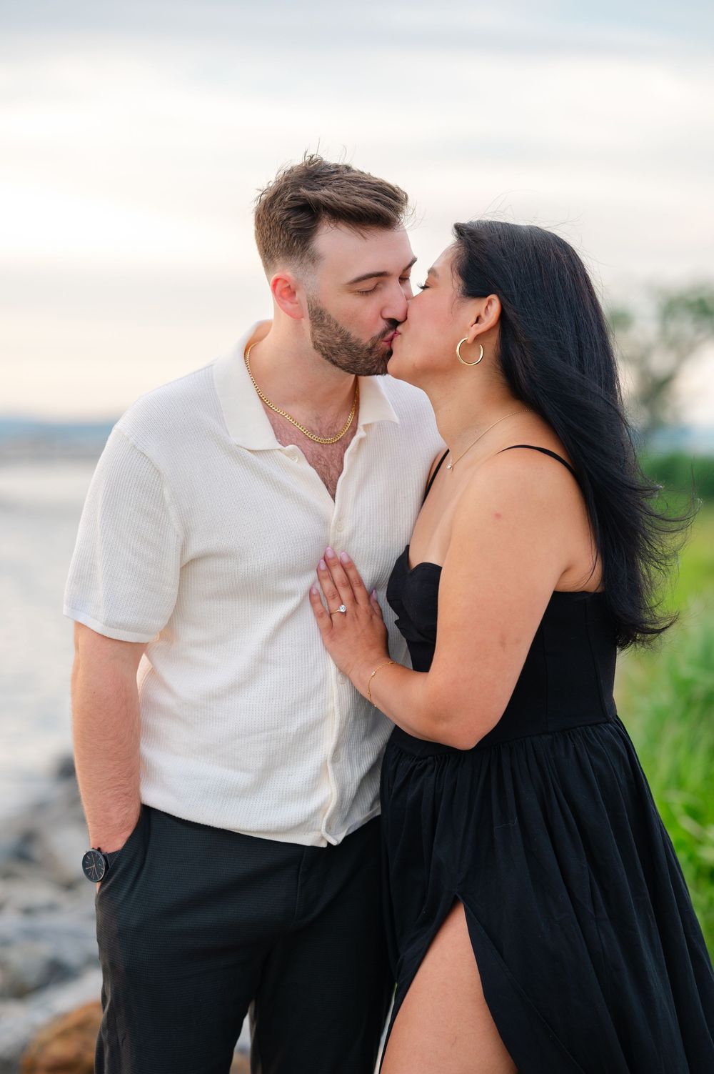 Couple sharing a romantic kiss near the waterfront, man in white shirt and woman in black dress holding hands.