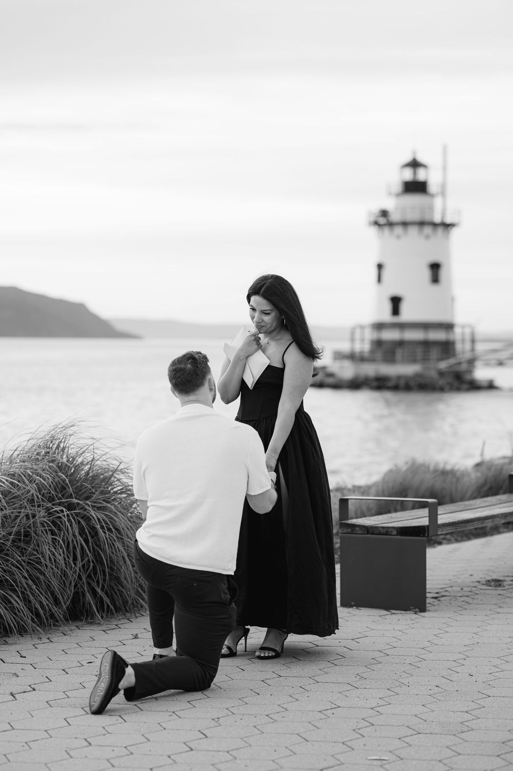 Black and white photo of a man on one knee proposing to a woman near a lighthouse by the water.
