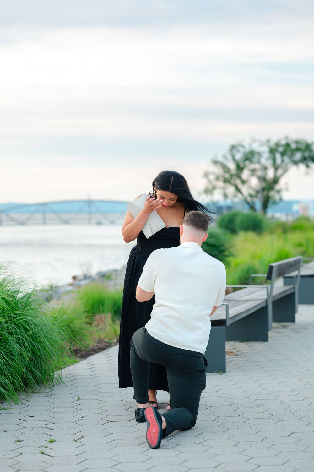 Emotional marriage proposal moment with man on one knee by scenic waterfront.