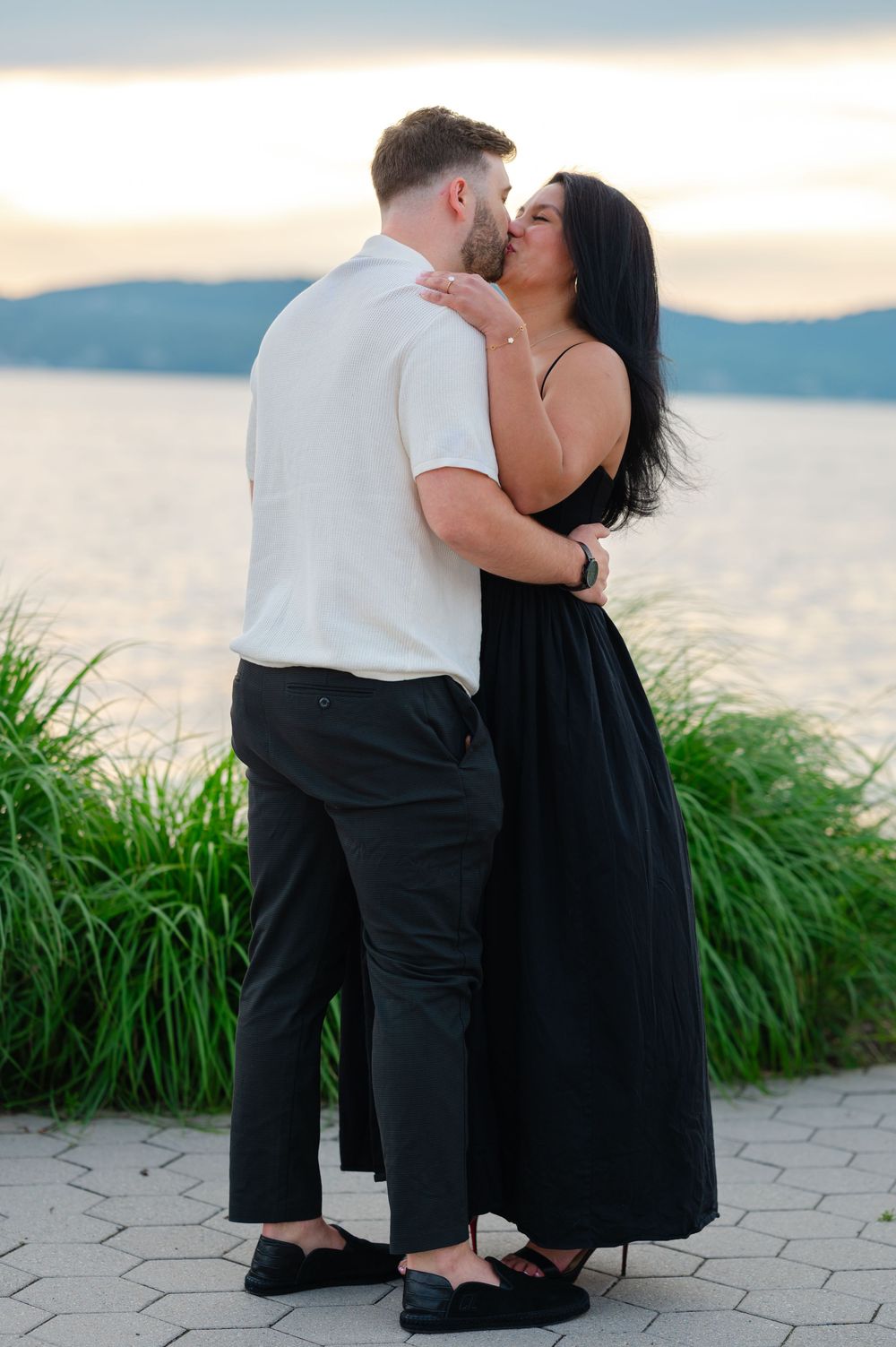 Couple sharing a romantic kiss at sunset by the waterfront, woman in black dress embracing man in white shirt.