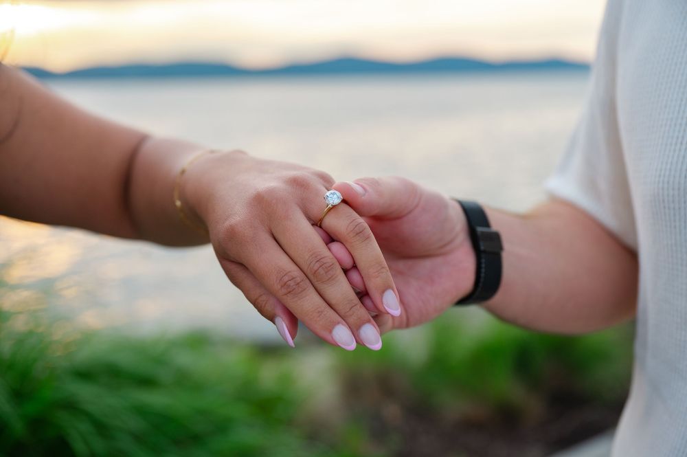 Close-up of couple holding hands showing diamond engagement ring at sunset near water.