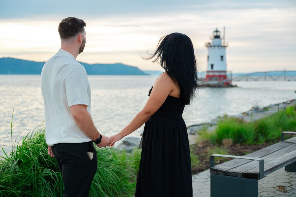 Couple holding hands facing a red and white lighthouse on the Hudson River at sunset.
