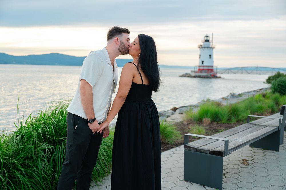 Romantic couple kissing while holding hands near a lighthouse by the water at dusk.
