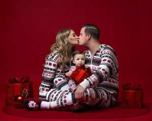 Family in matching Christmas pajamas with baby opening gift, surrounded by presents on red background.