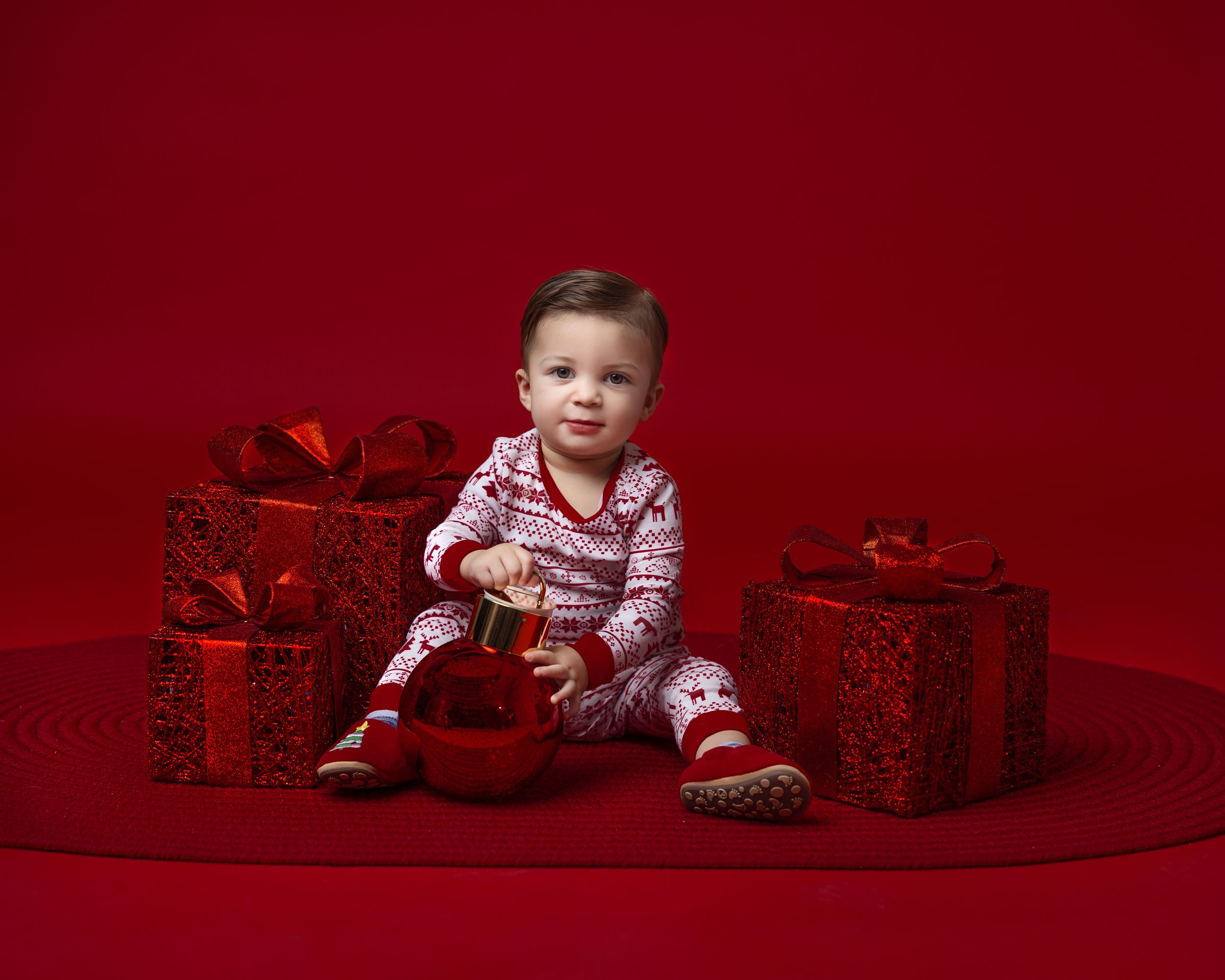 Baby in red Christmas pajamas sits surrounded by wrapped red gifts on a red background.