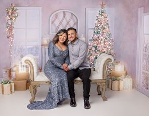 Couple in elegant attire sits together smiling beside a pink and white decorated Christmas tree.