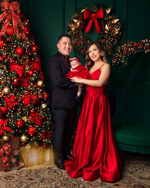Family Christmas portrait with man in black suit, woman in red gown holding baby, beside decorated tree and wreath.