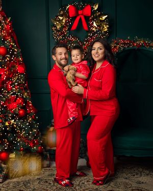 Family of three in matching red Christmas pajamas posing by decorated tree and wreath for holiday photo.