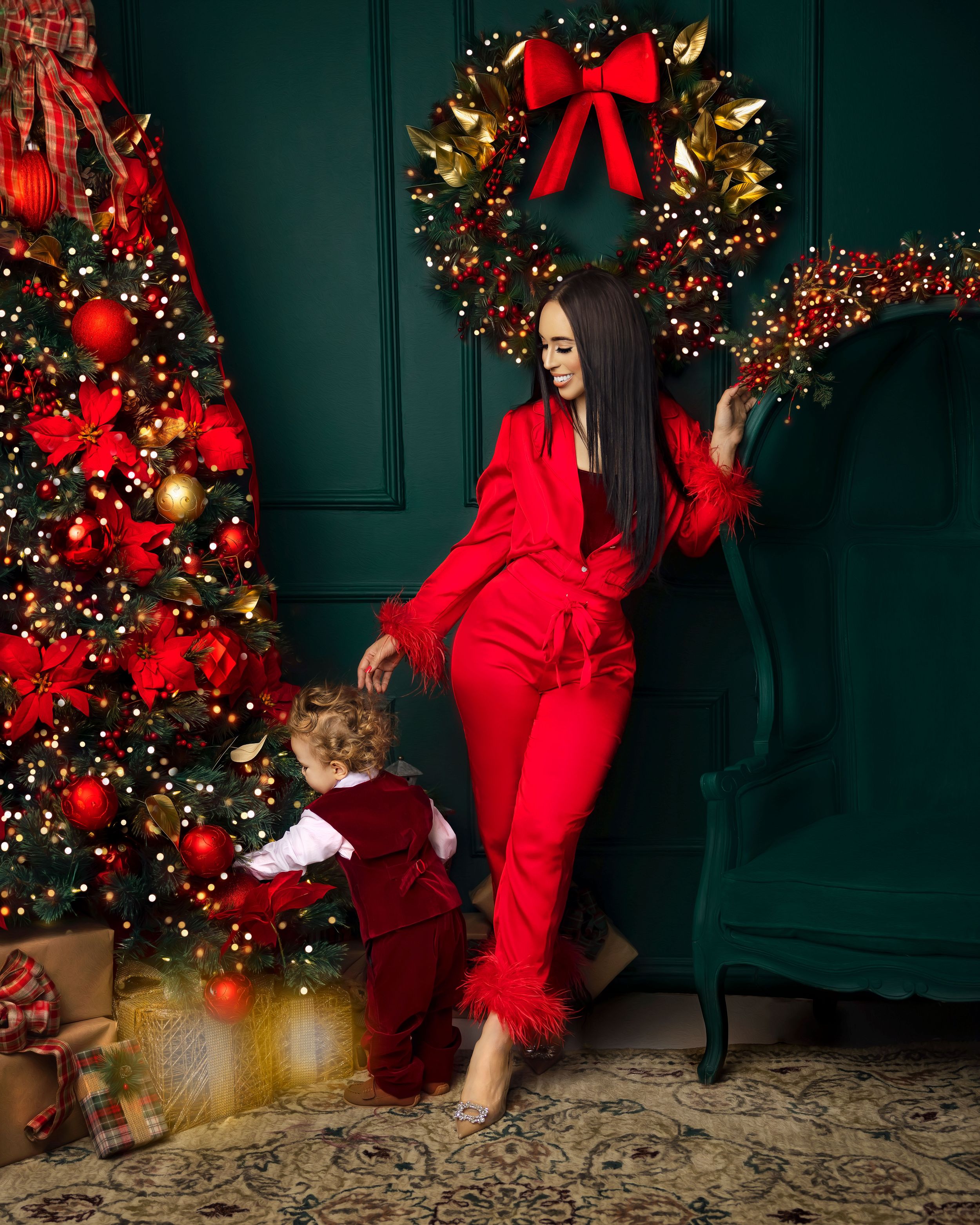 Woman in red feathered suit poses with toddler by Christmas tree and wreath.