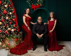 Family of three posing for a festive Christmas portrait with a decorated tree, wearing elegant red and black holiday outfits.