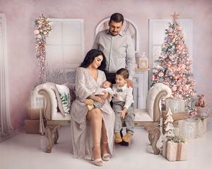 Family Christmas portrait with mother holding newborn, father, and toddler son beside a decorated pink Christmas tree.