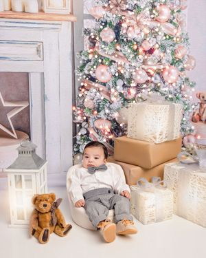 Baby in bow tie sits among Christmas gifts and teddy bear before a decorated pink and white holiday tree.