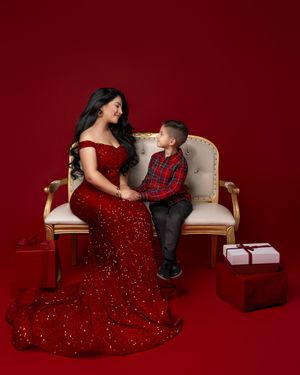 Woman in red sequin gown sits with young boy on elegant settee, surrounded by gift boxes on red background.