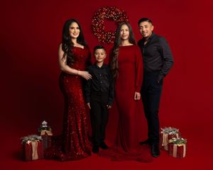 Family of four in matching red outfits posing for a Christmas portrait with gifts and wreath on red background.