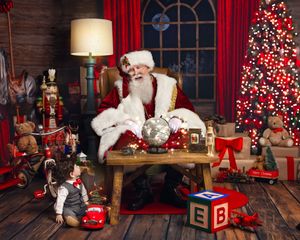 Santa Claus sits at a wooden table in a festive room with a lit Christmas tree, toys, and wrapped gifts around him.