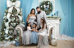 Family of five poses in silver-blue holiday attire beside decorated Christmas tree and wreath in festive studio setting.