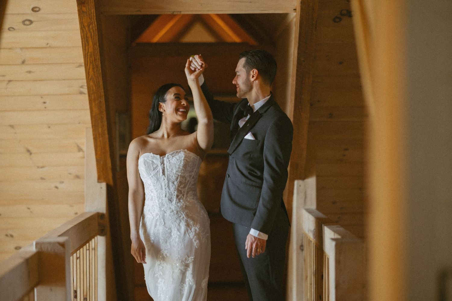 Bride in lace off-shoulder wedding gown holding champagne in rustic cabin kitchen with groom in tuxedo behind her.