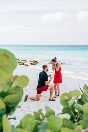 Man proposing on one knee to woman in red dress on tropical white sand beach with turquoise ocean backdrop.