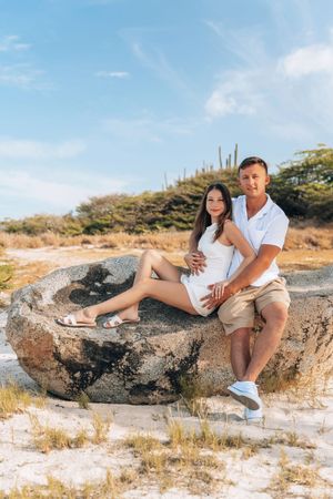 Couple in white outfits sitting on a large rock at a sandy beach with cacti and blue sky in Aruba.