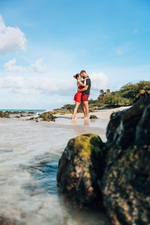 Couple embracing romantically on a tropical beach with rocks, turquoise water, and blue sky in the background.