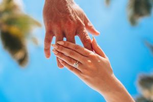 Two hands touching outdoors, woman wearing a diamond engagement ring against a bright blue sky with palm trees.