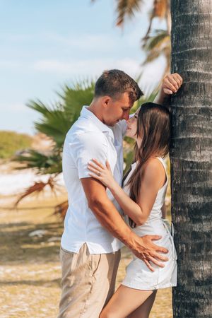 Romantic couple embracing near a palm tree on a tropical beach, wearing white outfits on a sunny day.
