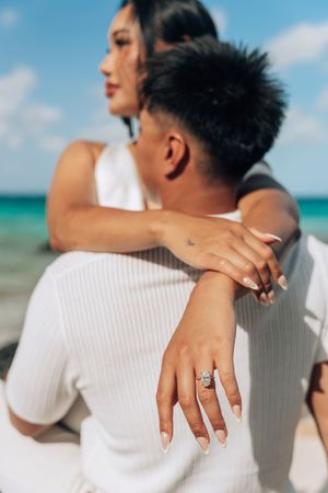 Woman showing off diamond engagement ring while embracing man from behind on a sunny beach.