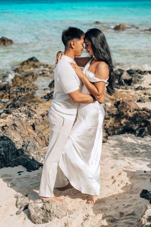 Couple in white outfits embracing on a sandy beach with turquoise ocean water and rocks in background.