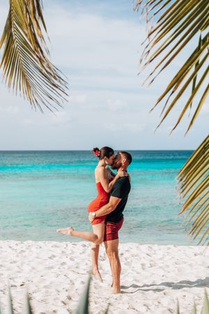 Romantic couple kissing on a white sand tropical beach with turquoise ocean water and palm tree fronds framing the scene.