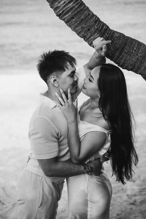 Black and white photo of romantic couple kissing near a palm tree on a tropical beach.