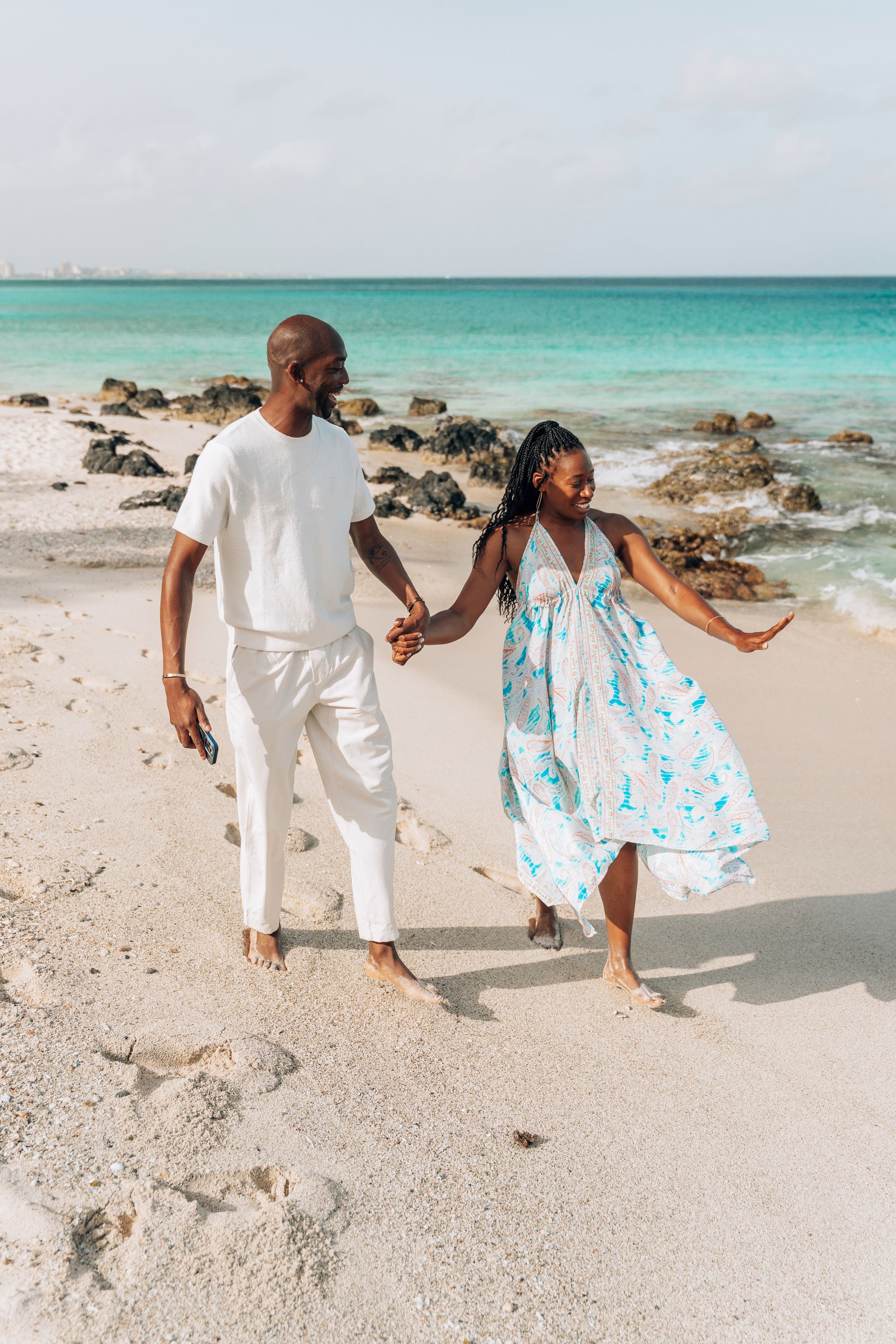 Happy couple holding hands strolling along a sunny Caribbean beach with turquoise waves and rocky shore.