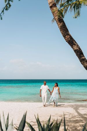 Couple holding hands walking on a tropical white sand beach with turquoise water and a palm tree.