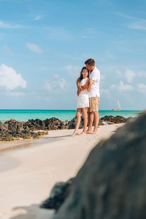 Romantic couple embracing on a white sandy tropical beach with turquoise ocean and sailboat in the background.