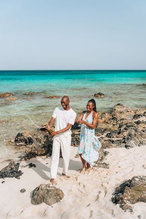 A joyful couple celebrating on a tropical beach with champagne, wearing white and floral outfits against turquoise waters.