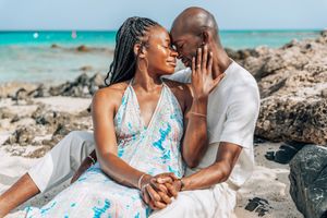 A romantic Black couple shares an intimate moment on a rocky beach, holding hands in colorful summer attire.