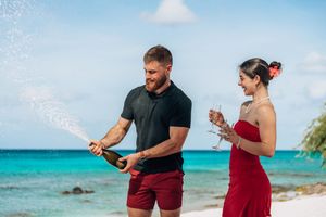 Couple celebrating on tropical beach, man popping champagne bottle with spray, woman in red dress holding glass.
