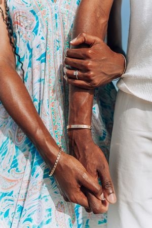 Close-up of a couple holding hands, wearing gold bracelets and an engagement ring, dressed in white and blue patterned outfits.
