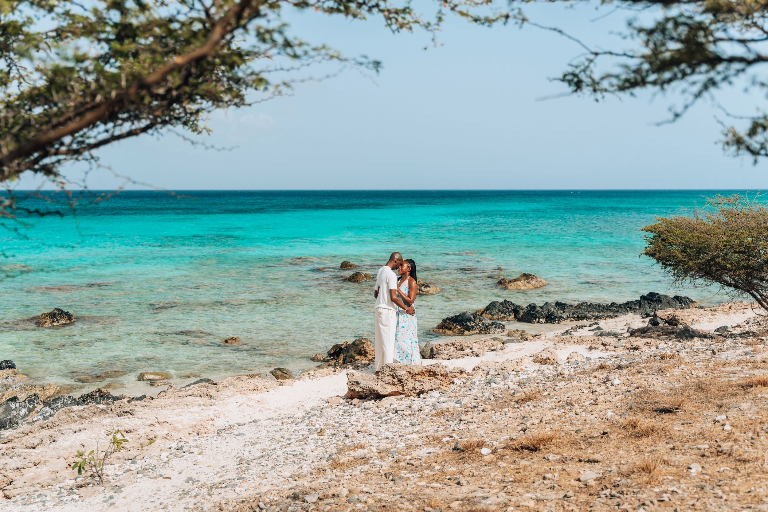 Couple in white embracing on rocky Caribbean beach with turquoise water and tree canopy in Aruba.