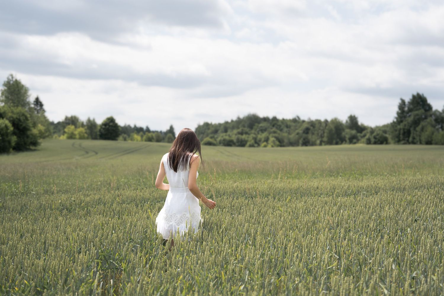 Woman in white dress walking through lush green field with trees in background under cloudy sky.