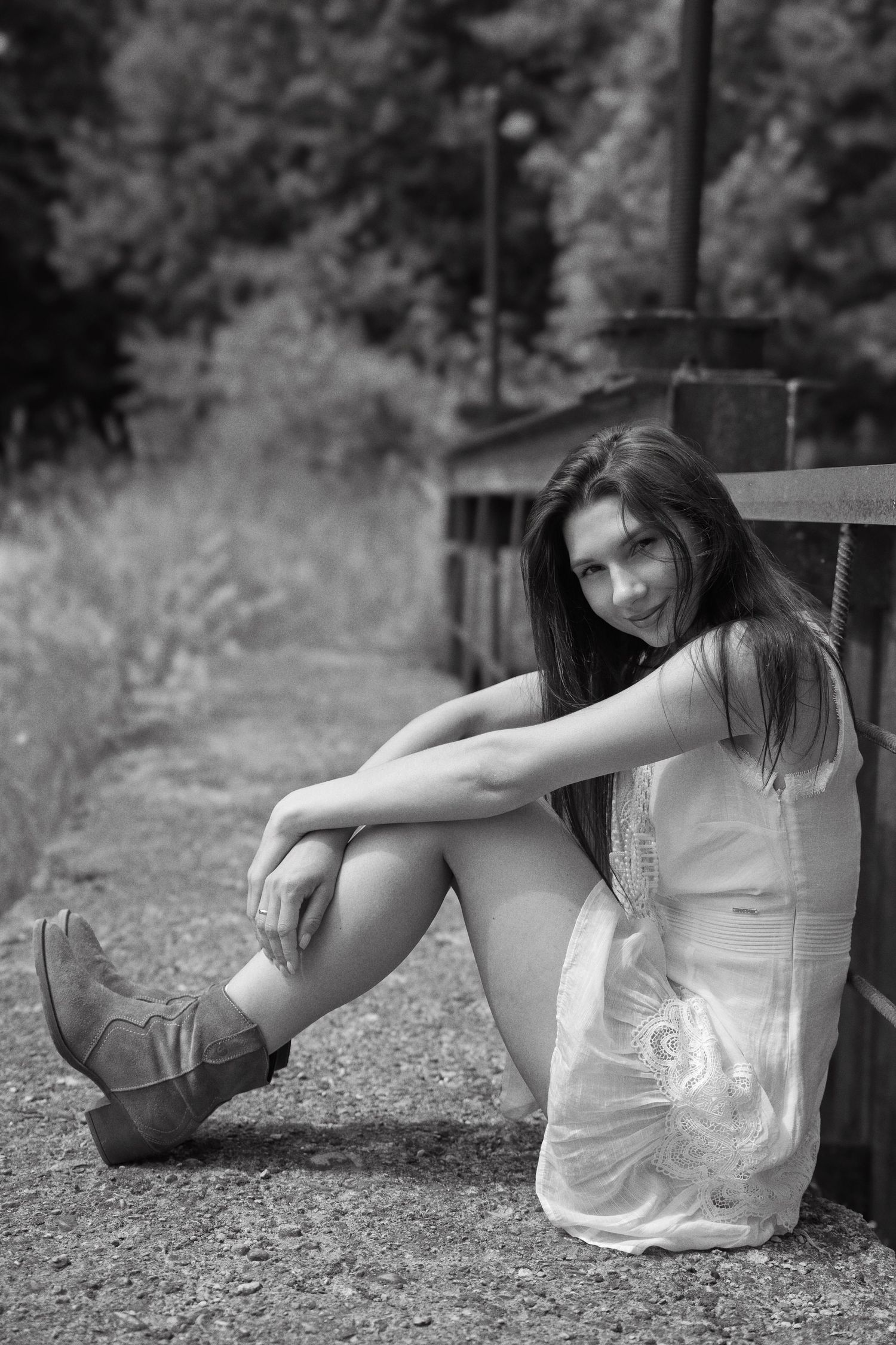 Young woman in white dress and cowboy boots sitting against wooden fence in black and white outdoor photo.