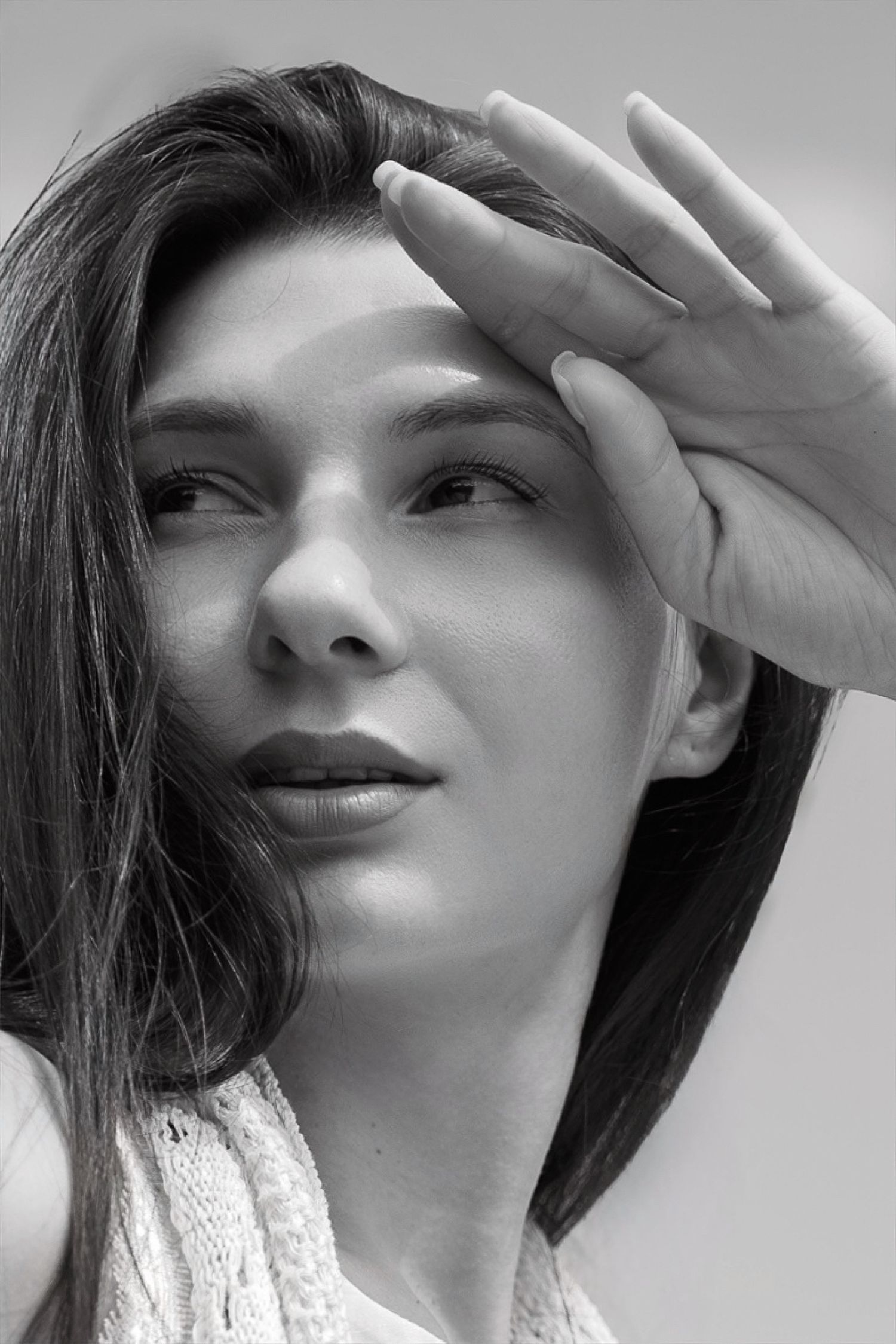 Black and white portrait of a young woman shielding her eyes with her hand, looking upward thoughtfully.