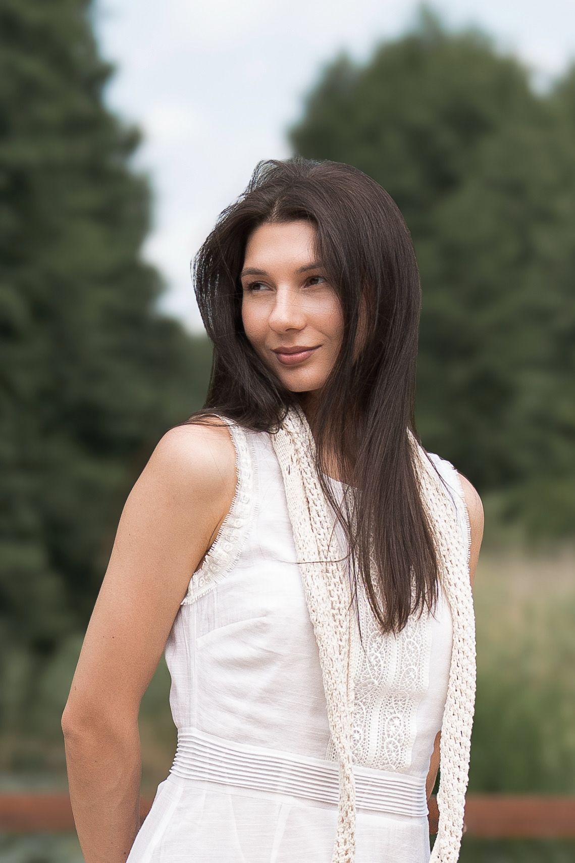 A young woman with long dark hair wearing a white sleeveless dress stands outdoors with green trees in the background.