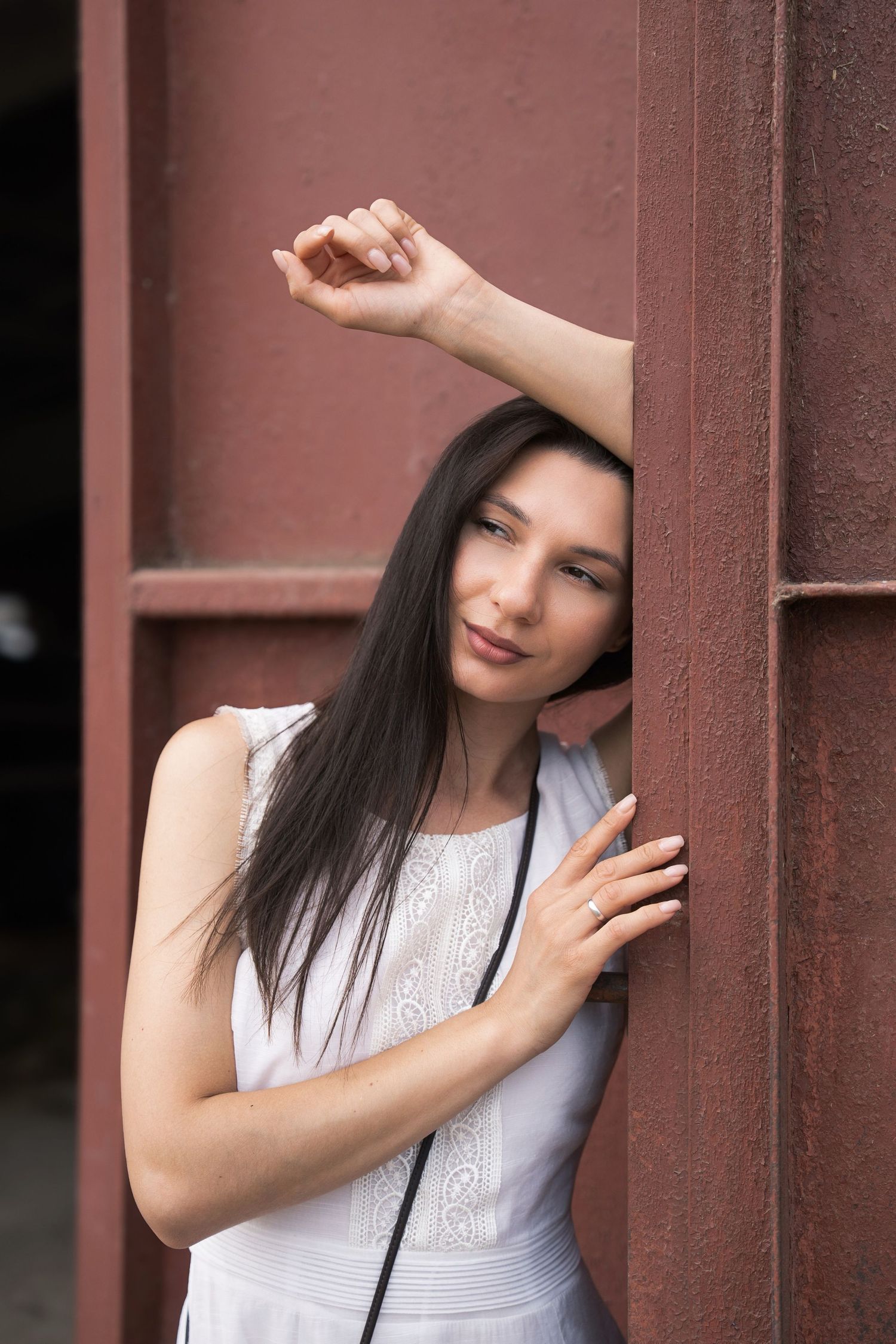 Brunette woman in white dress leaning against a rusty red metal wall, posing gracefully.