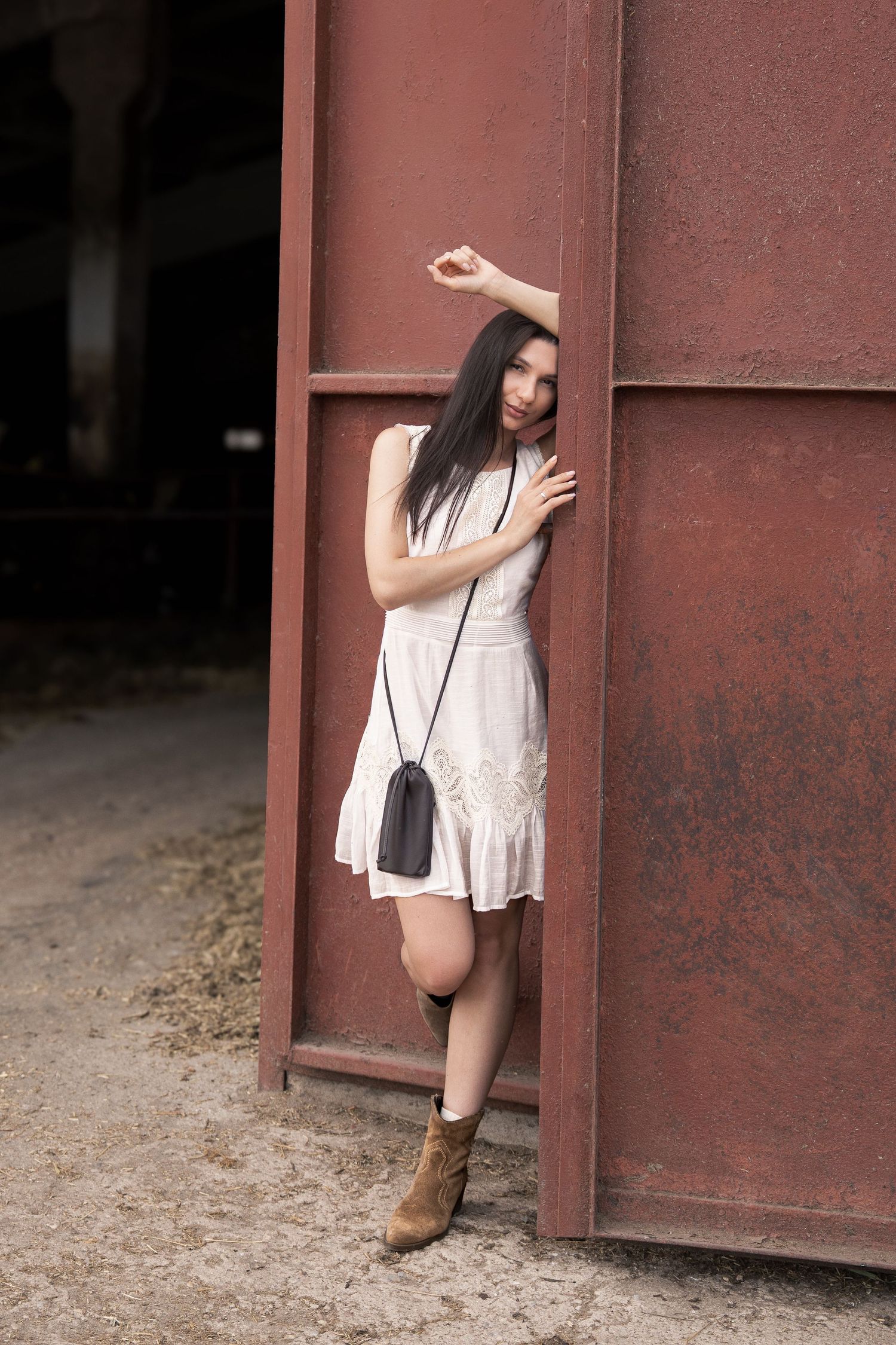 Young woman in white dress and brown boots poses against rusty red metal wall with small black crossbody bag.